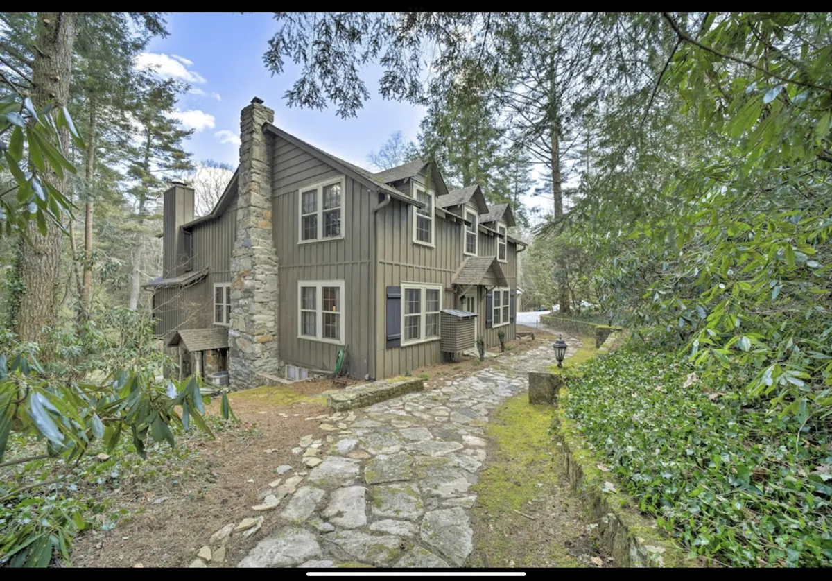 A welcoming exterior view of the property featuring a stone chimney, wooden siding, multiple large windows, and a beautifully landscaped pathway leading toward the entrance. The scene is set with lush garden plantings and mature trees, evoking an inviting atmosphere that connects the home to its natural surroundings.