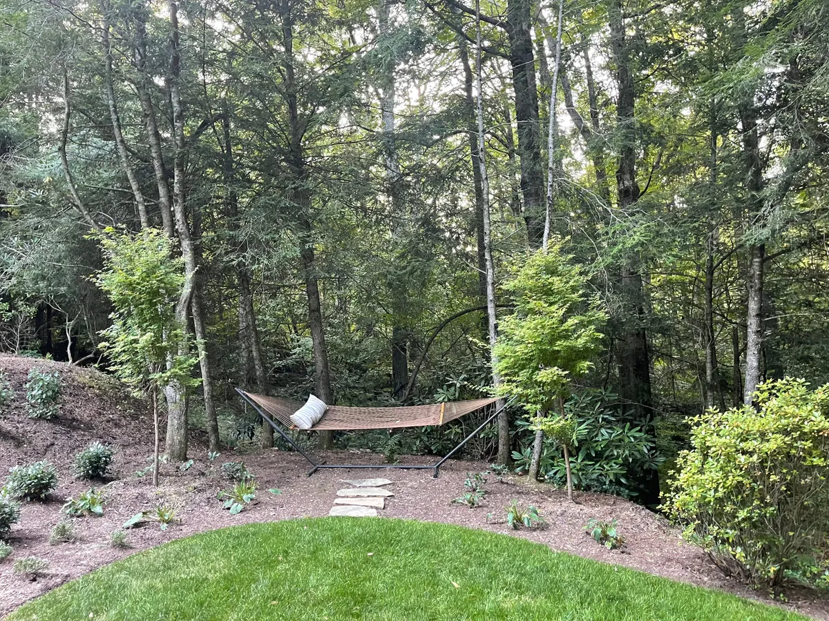 A hammock is strung between trees along a stone pathway, surrounded by a peaceful, wooded landscape. The area is shaded and quiet, with filtered sunlight creating a relaxing environment ideal for napping, reading, or unwinding.