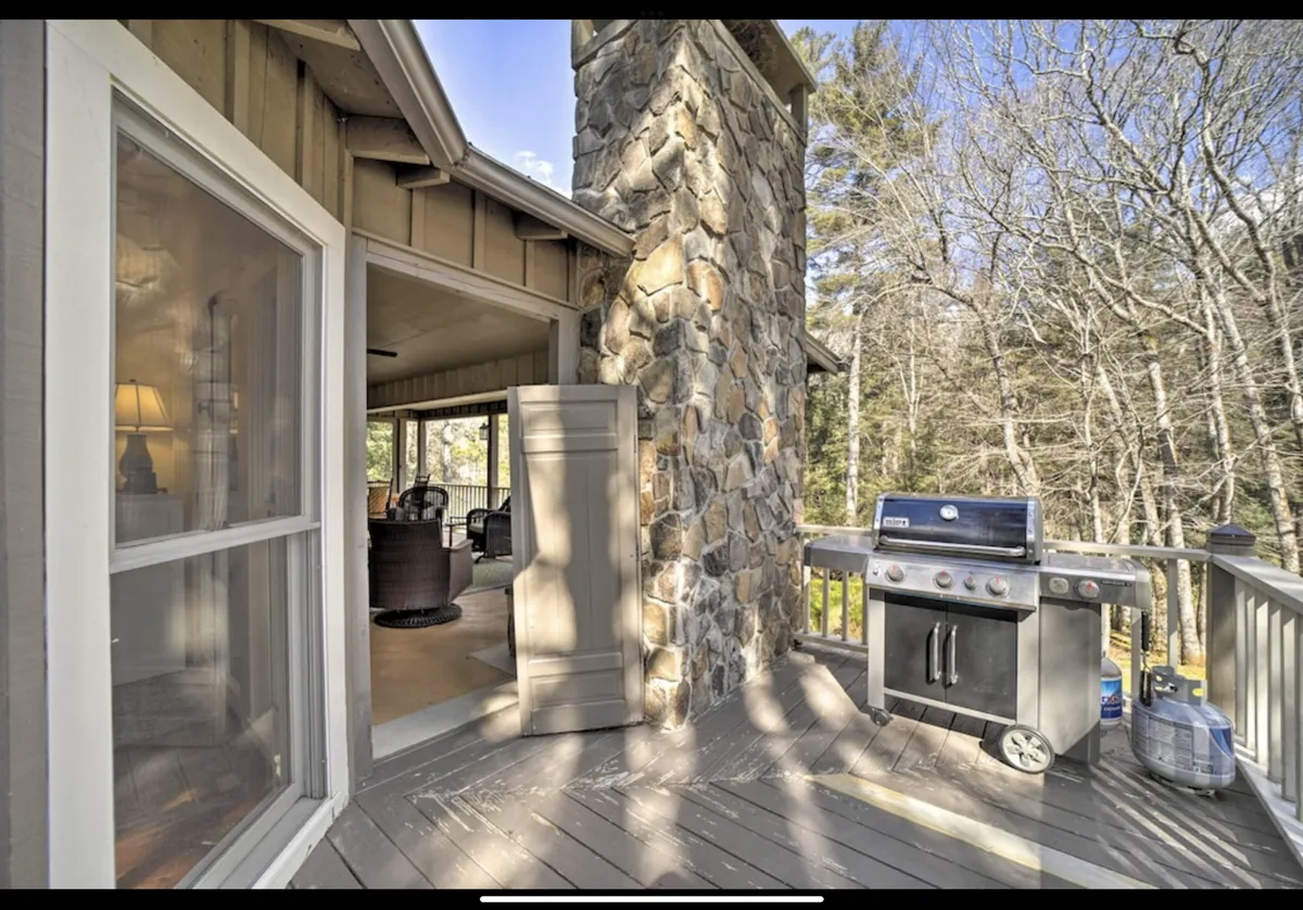 A cozy wooden deck with a stone chimney, built-in grill, and seating area. The deck is surrounded by lush greenery, offering a tranquil spot for outdoor dining or morning coffee. Warm, inviting lighting suggests the perfect setting for gatherings or quiet reflection.