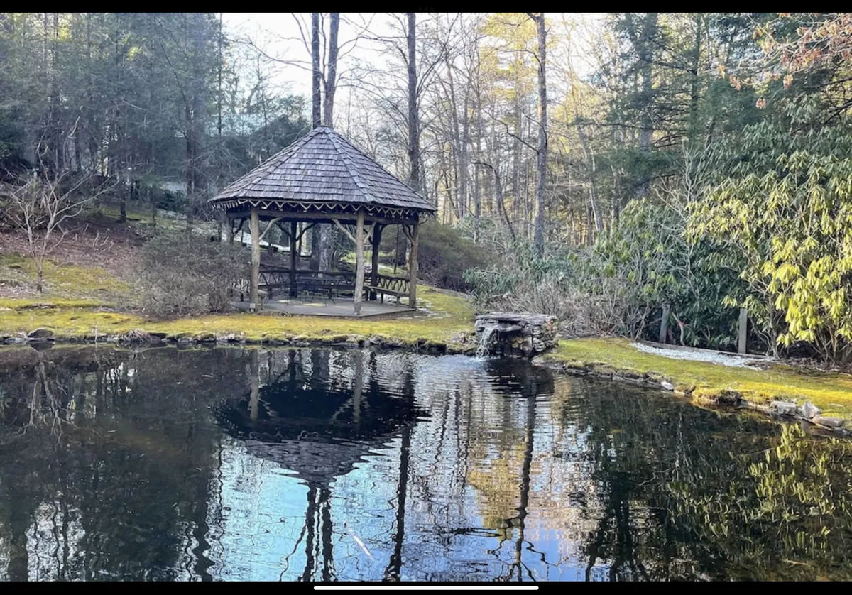 A picturesque gazebo overlooking a reflective pond, framed by mature trees. The setting is peaceful, with dappled sunlight and clear views of the water, inviting the viewer to imagine relaxing, reading, or meditating in the serene space.