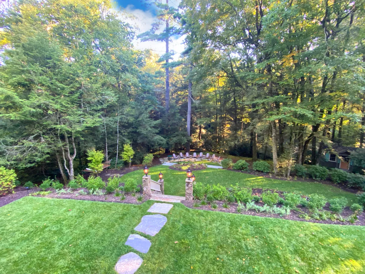 An inviting outdoor area centered around a stone pathway and a fire pit seating area, surrounded by lush landscaped gardens and dense trees. The space is designed for relaxing, gathering for intimate conversations, or celebrating special moments under the stars.
