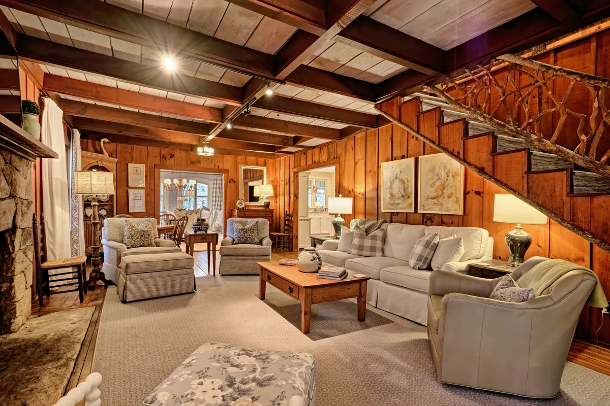The image captures a warm, inviting great room featuring a striking stone fireplace as its centerpiece, a plush and comfortable seating arrangement, and tasteful artwork adorning the walls. The open layout and visible wooden staircase enhance the sense of spaciousness and connection between areas.