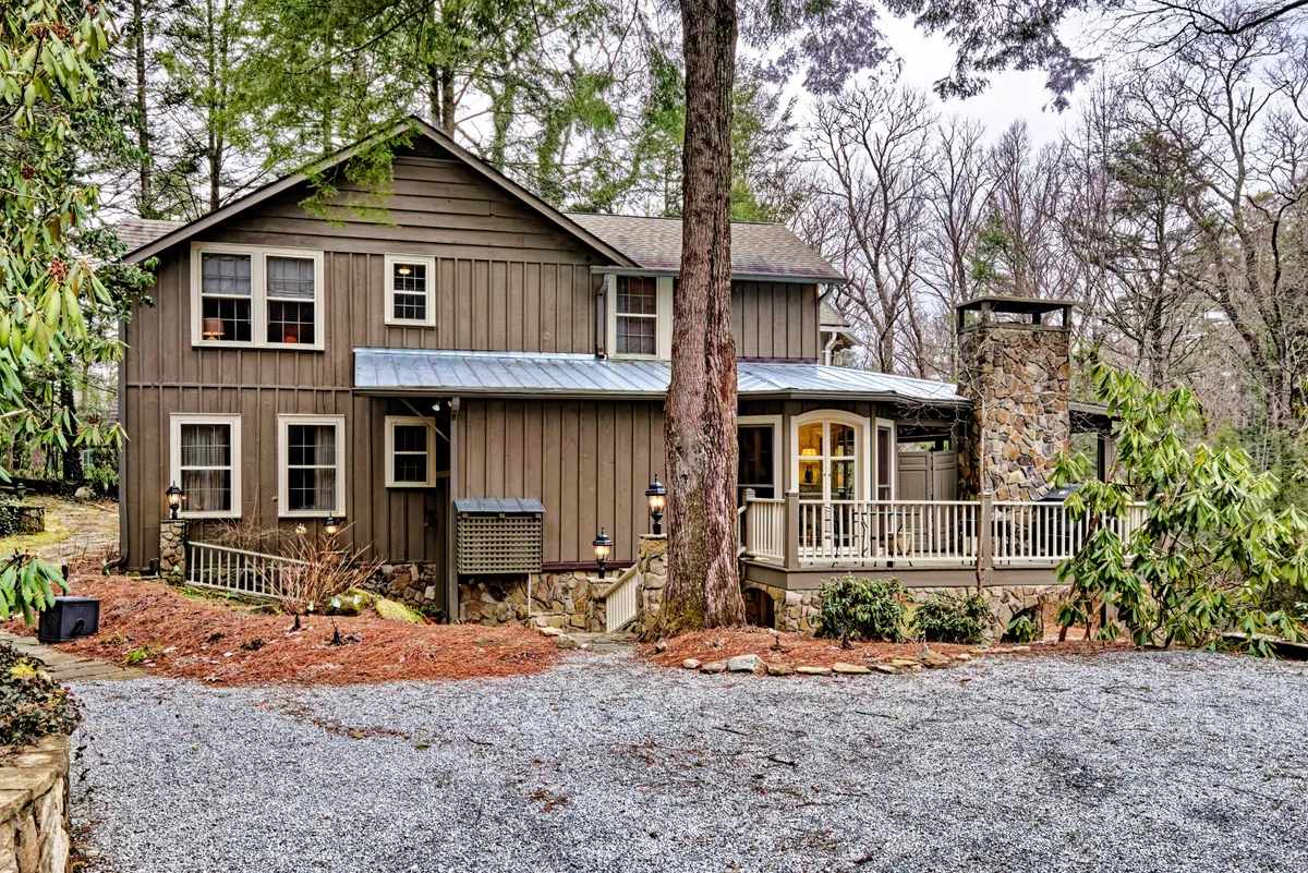 This image captures the home's exterior with a welcoming stone chimney, a cozy front porch, a gravel driveway, and surrounding trees. The environment feels tranquil and inviting, offering a glimpse into the natural setting that encourages outdoor gatherings and relaxation.