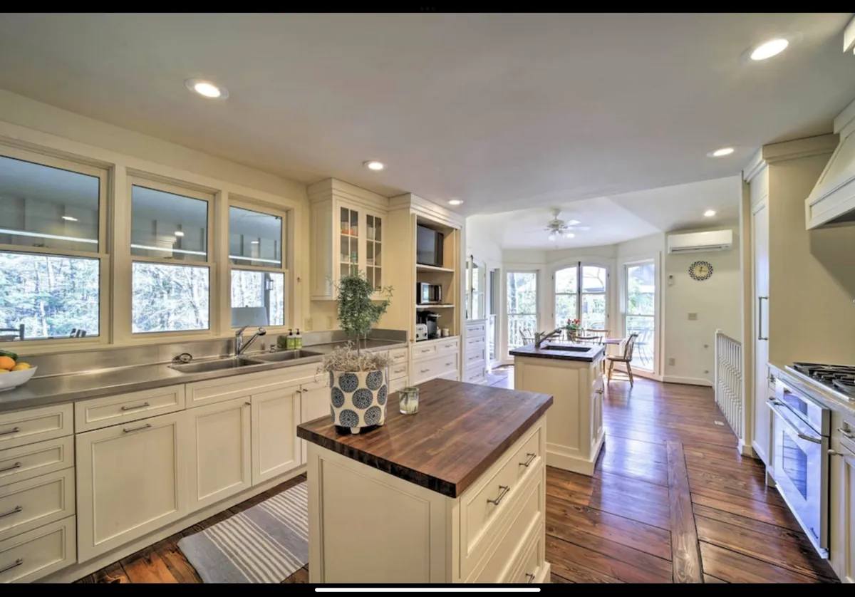 A chef-inspired kitchen centers around a large island, surrounded by stainless steel appliances, wood flooring, and ample natural light pouring in through a window. The space feels functional yet stylish, ready for social cooking and entertaining.