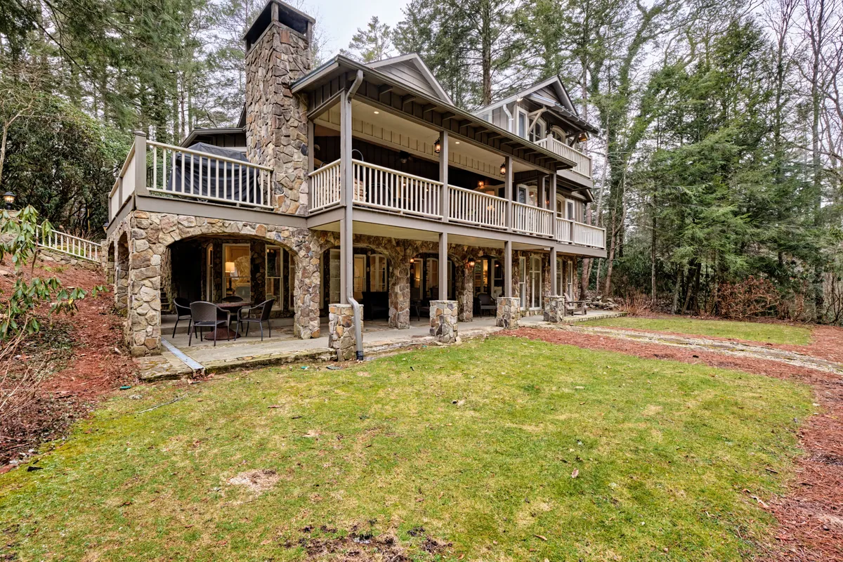 An expansive exterior shot showcasing a stone chimney, a large balcony perfect for outdoor dining or relaxation, a well-maintained lawn, and mature surrounding trees. The scene feels both tranquil and social, with spaces for group gatherings and moments of reflection.