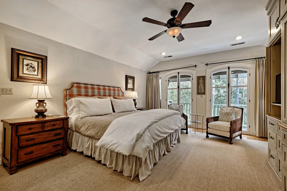 A tranquil bedroom featuring a bed with a patterned headboard, two bedside lamps, and an armchair positioned by a large window. Double doors open to the outdoors, allowing natural light to flood the space and creating a strong visual connection to nature.