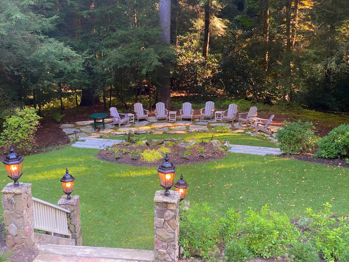 Set in a landscaped garden area, this image shows wooden Adirondack chairs arranged along a stone pathway, with lampposts and a lush garden as the backdrop. The scene evokes communal gatherings, quiet contemplation, and the enjoyment of natural surroundings.