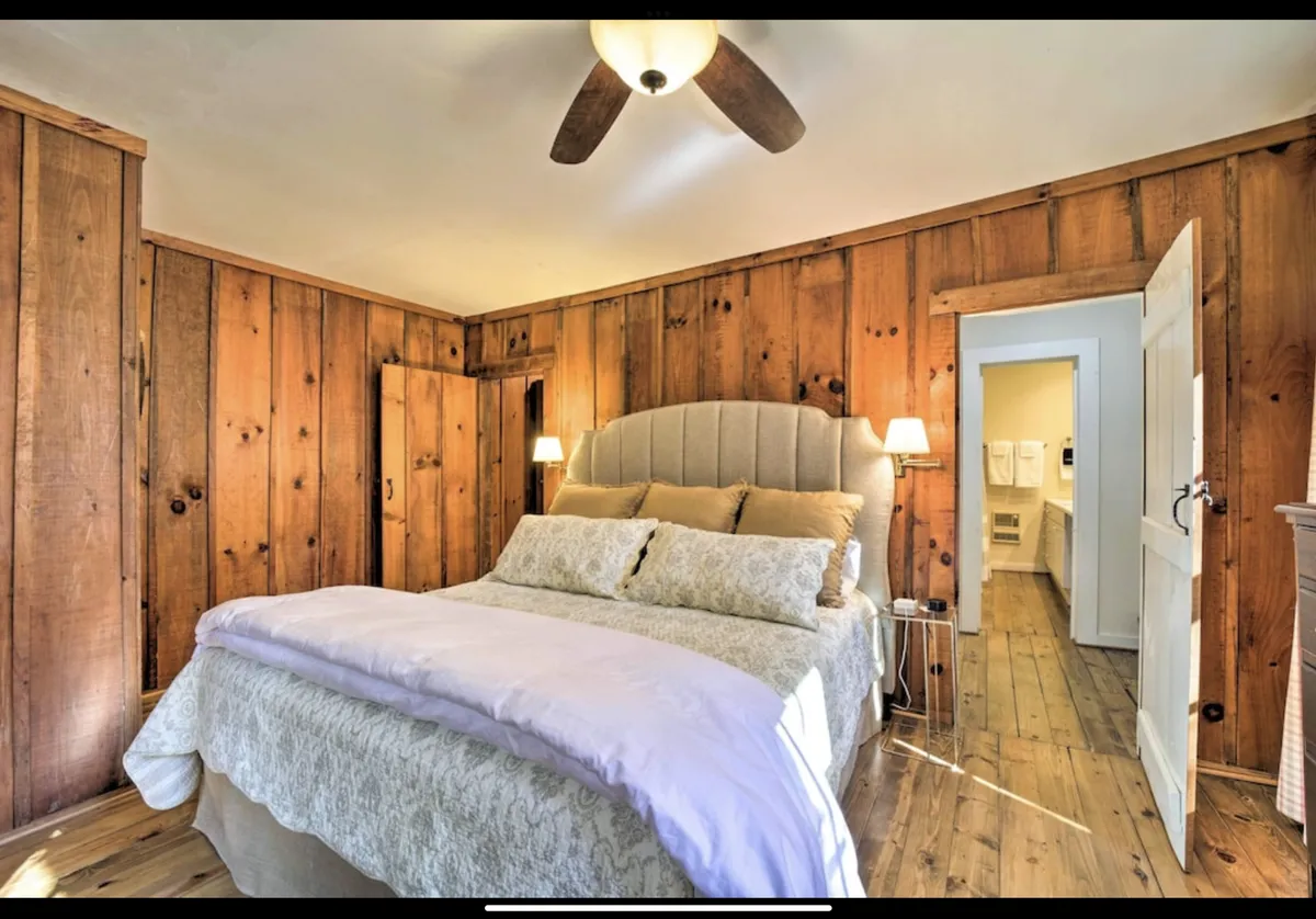 A welcoming bedroom with wood paneling, a bed flanked by nightstands, and a visible doorway leading directly to a bathroom. The space exudes warmth and privacy, emphasizing the en-suite feature.