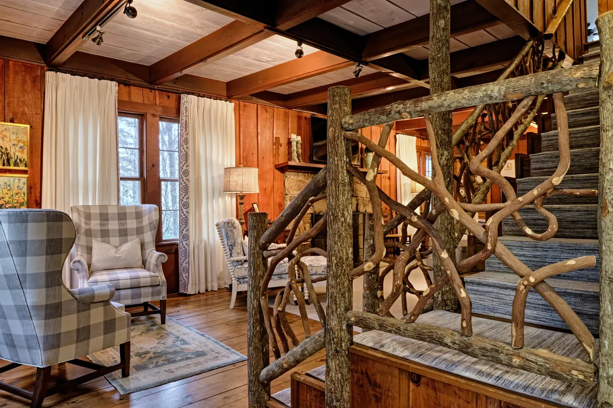 A rustic interior view featuring a plaid armchair beside a wooden staircase, exposed wooden beams on the ceiling, and soft lamplight creating a warm, inviting glow. The scene suggests a welcoming retreat after exploring outdoors.