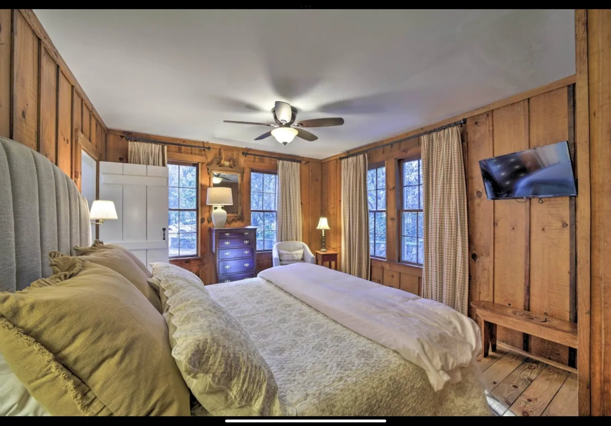A cozy bedroom featuring a comfortable bed with decorative pillows, warm wooden walls, windows adorned with curtains, a chest of drawers, and a flat-screen TV. Gentle natural light fills the room, creating an atmosphere of comfort and tranquility.